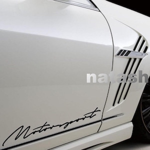 Op de afbeelding: Witte auto met zwarte details, waaronder een "Motorsport" sticker en een zwarte velg. De auto heeft een sportief design met een ventilatieopening en verticale zwarte strepen.