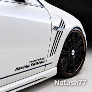 Op de afbeelding: Een wit zijpaneel van een auto met een zwarte sticker met de tekst Powered by Racing Edition. Het voertuig is voorzien van een verchroomde zijventilatie en een groot zwart meerspaaks wiel met een bronskleurige velgrand en een Wald-logo.