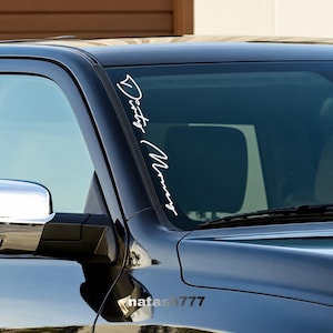 May include: A white vinyl decal applied to the passenger side of a dark vehicle windshield. The decal features the text Dirty Money written in a stylized, cursive script font oriented vertically along the edge of the glass.