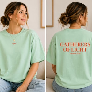 May include: A pale green t-shirt with the word "light" in red on the front. The back of the shirt displays the text "Gatherers of Light" and "Romans 10:14" in red. The t-shirt has a loose fit.