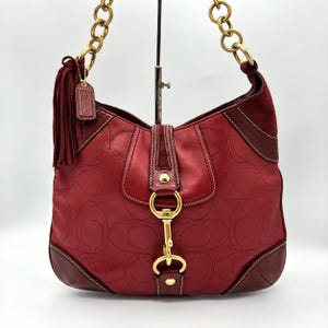 May include: A deep red shoulder bag featuring a debossed monogram pattern, a gold-tone chain strap, and a central gold-tone metal clasp closure. A matching red suede tassel hangs from the side, and the bag includes smooth leather trim.