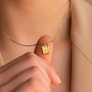 Puede incluir: Un delicado collar de oro con dos pequeños colgantes rectangulares. Los colgantes están grabados con las letras "K" y "T". El collar se muestra en un dedo, destacando los finos detalles y el diseño elegante de la joya.