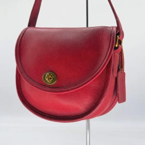 May include: A red leather saddle bag featuring a curved flap, a gold-tone metal turn-lock closure, and a matching shoulder strap. A rectangular leather hang tag is attached to the side of the bag with a gold-tone metal chain.