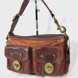 May include: A brown leather shoulder bag featuring two front exterior pockets with brass turn-lock closures, a top zipper, and a detachable shoulder strap with brass hardware. The bag has visible stitching details and a weathered, textured finish.