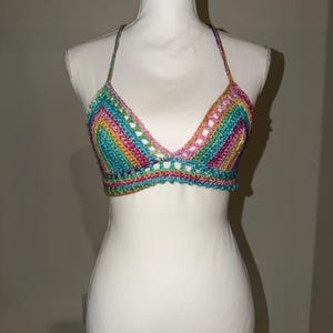 May include: A handmade, colorful crochet top displayed on a white mannequin. The top features a rainbow of colors, including pink, blue, yellow, and green, in a striped pattern. The halter top has a triangle shape with a scalloped edge.