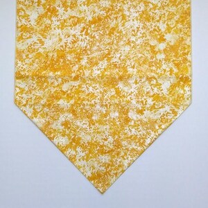 May include: A yellow table runner with a white splatter pattern. The runner is made of fabric and has a pointed end.