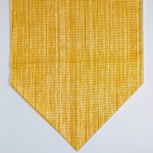 May include: A golden yellow table runner with a textured, woven appearance. The runner has a pointed end and is set against a white background. The fabric has a subtle, grid-like pattern.