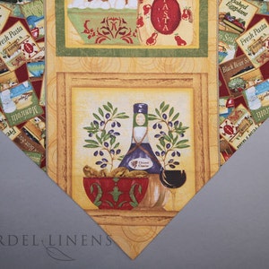 Puede incluir: Un camino de mesa de tela amarillo y marrón con un patrón repetido de etiquetas de comida y bebida italianas. El centro del camino de mesa presenta una ilustración enmarcada de una botella de aceite de oliva, un tazón de aceitunas y una rama de olivo.