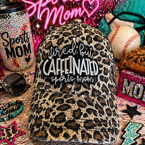 Puede incluir: Gorra de béisbol con estampado de leopardo y el texto "tired but caffeinated sports mom". La imagen incluye accesorios deportivos, como un vaso adornado con pedrería y pelotas de béisbol.