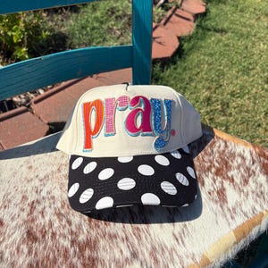 Puede incluir: Una gorra de béisbol de color crema con la palabra pray en letras coloridas, texturizadas y con varios patrones en el panel frontal. La visera es negra con un patrón repetido de lunares blancos y la gorra tiene una corona estructurada.
