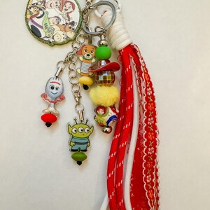 May include: A keychain featuring a circular charm with Toy Story characters, a Forky charm, an alien charm, and a Slinky Dog charm. It includes a beaded strand with a red hat, a yellow pom-pom, and a red and white fabric tassel with a silver clasp.