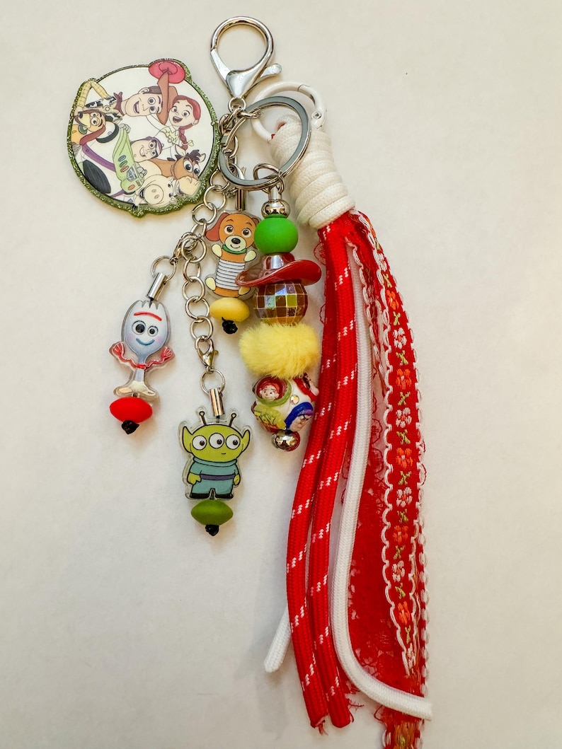 May include: A keychain featuring a circular charm with Toy Story characters, a Forky charm, an alien charm, and a Slinky Dog charm. It includes a beaded strand with a red hat, a yellow pom-pom, and a red and white fabric tassel with a silver clasp.