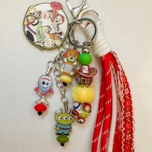 May include: A keychain featuring a circular charm with Toy Story characters, a Forky charm, an Alien charm, and a Slinky Dog charm. It includes a beaded strand with a disco ball bead, a yellow pom-pom, and a red fabric tassel with white trim.