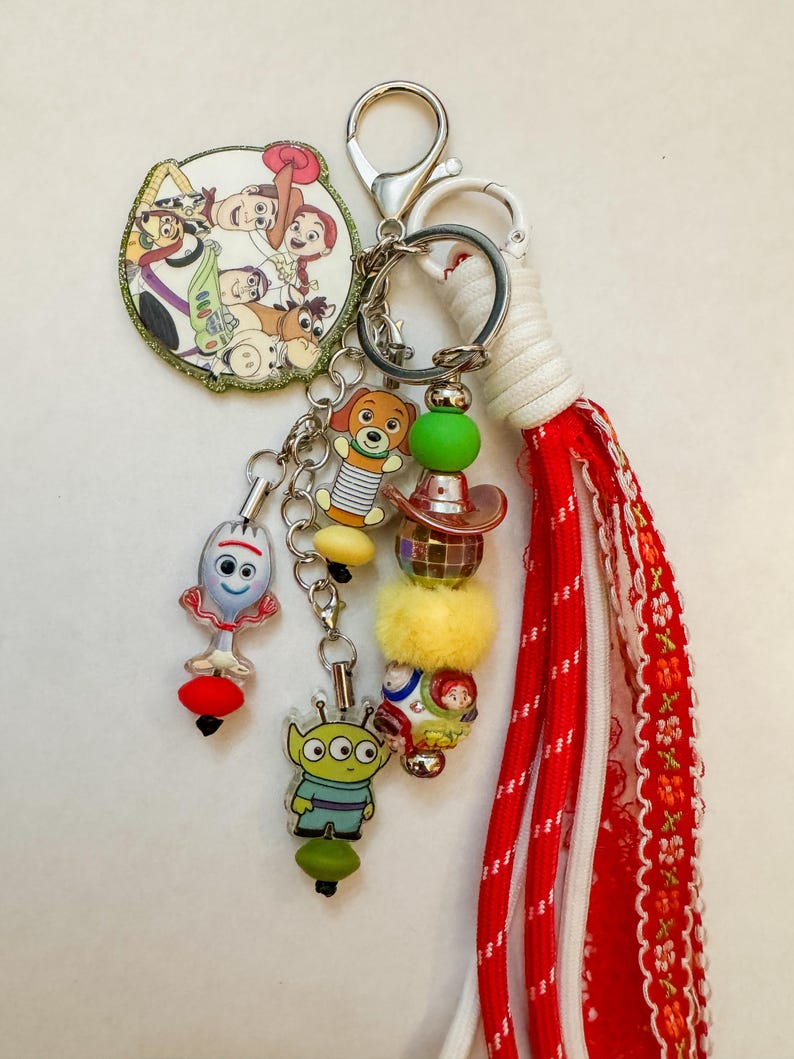 May include: A keychain featuring a circular charm with Toy Story characters, a Forky charm, an Alien charm, and a Slinky Dog charm. It includes a beaded strand with a disco ball bead, a yellow pom-pom, and a red fabric tassel with white trim.