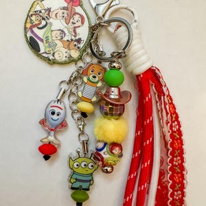 May include: A keychain featuring a circular charm with Toy Story characters, a Forky charm, an alien charm, a Slinky Dog charm, and a beaded strand with a disco ball and cowboy hat. A long red and white fabric tassel is attached to the silver metal rings.