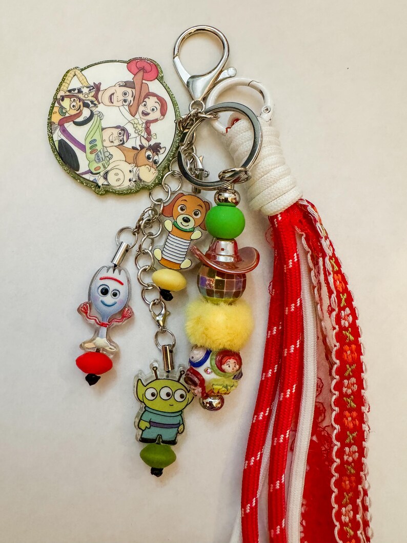 May include: A keychain featuring a circular charm with Toy Story characters, a Forky charm, an alien charm, a Slinky Dog charm, and a beaded strand with a disco ball and cowboy hat. A long red and white fabric tassel is attached to the silver metal rings.