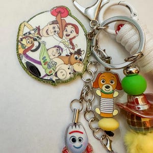 May include: A keychain featuring a circular charm with Toy Story characters including Woody, Jessie, Buzz Lightyear, Bullseye, and Hamm. Additional charms include a Slinky Dog, a Forky figure, and a colorful assortment of beads and a yellow pom-pom.
