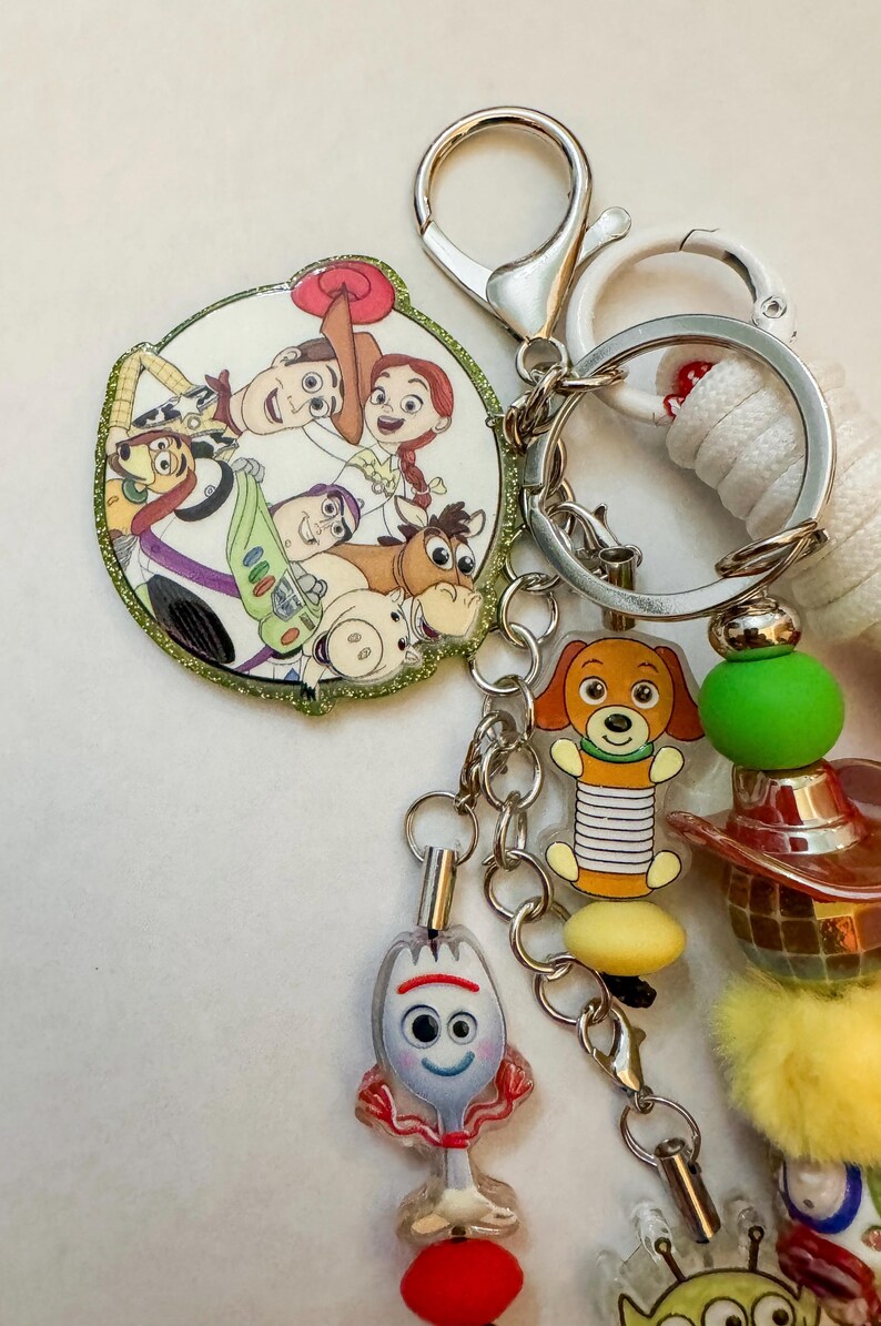 May include: A keychain featuring a circular charm with Toy Story characters including Woody, Jessie, Buzz Lightyear, Bullseye, and Hamm. Additional charms include a Slinky Dog, a Forky figure, and a colorful assortment of beads and a yellow pom-pom.