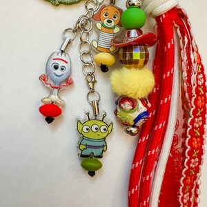 May include: A keychain featuring multiple charms including a white spork character, a green three-eyed alien, a brown dog, and a round bead with character graphics. It includes a yellow fuzzy bead, a disco ball bead, and a red fabric tassel.