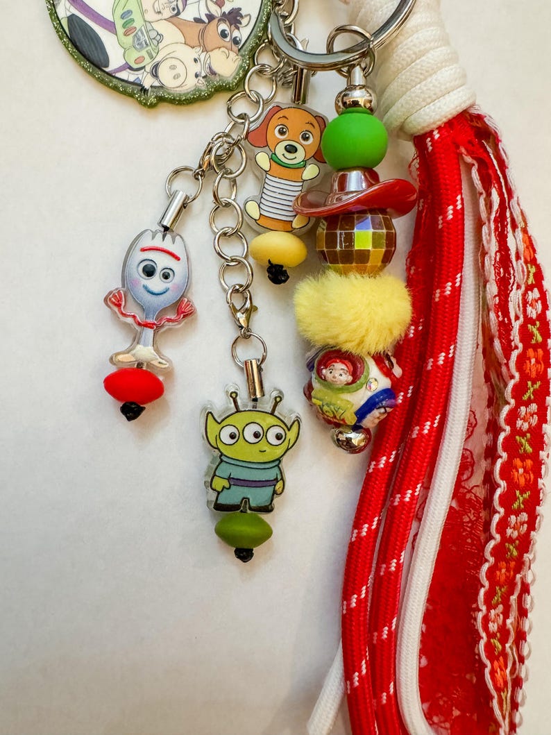 May include: A keychain featuring multiple charms including a white spork character, a green three-eyed alien, a brown dog, and a round bead with character graphics. It includes a yellow fuzzy bead, a disco ball bead, and a red fabric tassel.