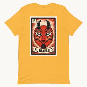 Peut inclure: Un t-shirt jaune moutarde avec une impression graphique rectangulaire représentant un visage de diable rouge avec des cornes noires, une moustache et un bouc. Le motif ressemble à une carte à jouer avec le chiffre 2 et le texte EL DIABLITO.