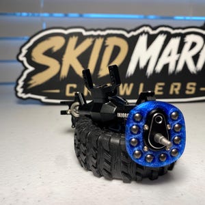 Op de afbeelding: Een zwarte RC-crawler-asconstructie met een blauwe glitterende wielnaaf, zilverkleurige hardware en een zwarte rubberen band met profiel. Het onderdeel staat voor een zwart-beige bord met de tekst SKID MARK CRAWLERS.