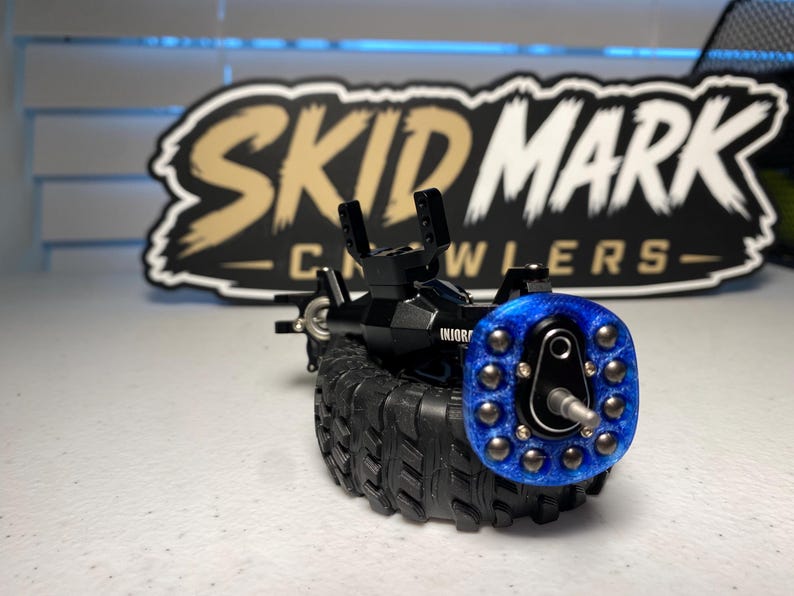 Op de afbeelding: Een zwarte RC-crawler-asconstructie met een blauwe glitterende wielnaaf, zilverkleurige hardware en een zwarte rubberen band met profiel. Het onderdeel staat voor een zwart-beige bord met de tekst SKID MARK CRAWLERS.