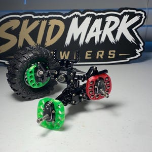 Op de afbeelding: Een zwarte metalen RC-crawler-asconstructie met &eacute;&eacute;n zwarte rubberen band, twee doorschijnende groene wielnaven en &eacute;&eacute;n doorschijnende rode wielnaaf. De constructie bevat zilverkleurige schroeven en metalen verbindingen. Bord met SKID MARK.