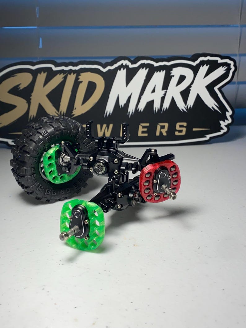 Op de afbeelding: Een zwarte metalen RC-crawler-asconstructie met &eacute;&eacute;n zwarte rubberen band, twee doorschijnende groene wielnaven en &eacute;&eacute;n doorschijnende rode wielnaaf. De constructie bevat zilverkleurige schroeven en metalen verbindingen. Bord met SKID MARK.