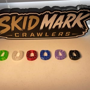 Op de afbeelding: Zes kleine, ringvormige fidget-speeltjes in groen, transparant, rood, blauw, paars en zwart, elk met ingebedde metalen kogellagers. Daarachter staat een zwart bord met witte en gouden tekst: SKID MARK CRAWLERS.