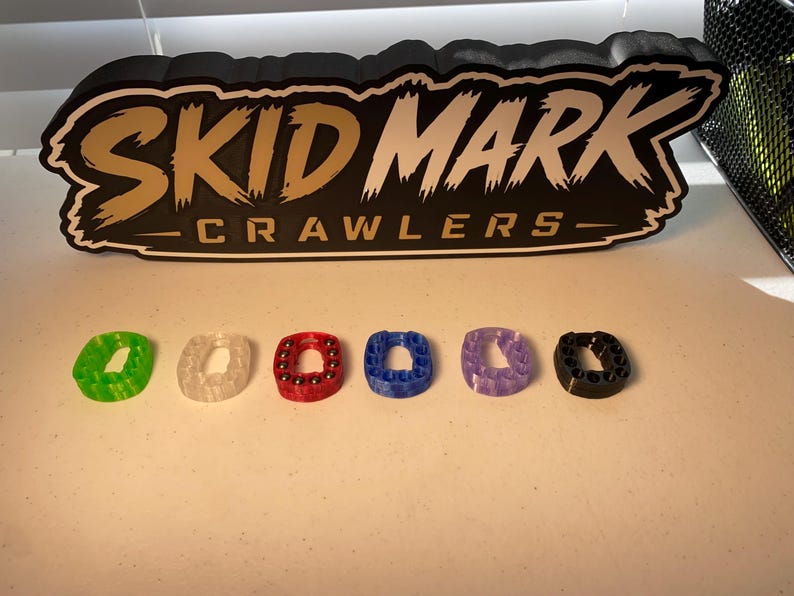 Op de afbeelding: Zes kleine, ringvormige fidget-speeltjes in groen, transparant, rood, blauw, paars en zwart, elk met ingebedde metalen kogellagers. Daarachter staat een zwart bord met witte en gouden tekst: SKID MARK CRAWLERS.