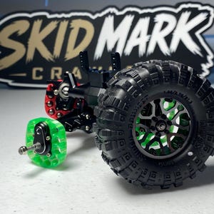 Op de afbeelding: Een zwart-groene asconstructie voor een op afstand bestuurbare crawler met een grote zwarte off-road band met diep profiel, een groene doorschijnende wielnaaf en een rood metalen onderdeel. Op de achtergrond staat een zwart bord met de tekst SKID MARK.