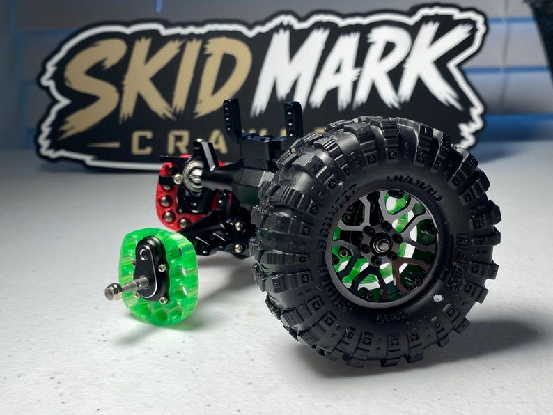 Op de afbeelding: Een zwart-groene asconstructie voor een op afstand bestuurbare crawler met een grote zwarte off-road band met diep profiel, een groene doorschijnende wielnaaf en een rood metalen onderdeel. Op de achtergrond staat een zwart bord met de tekst SKID MARK.