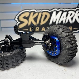 Op de afbeelding: Een zwarte metalen RC crawler as-assemblage met de merknaam INJORA in het wit gedrukt. De as is verbonden met twee zwarte rubberen banden met diep profiel, waarbij de rechterband op een felblauwe metalen wielnaaf is gemonteerd.
