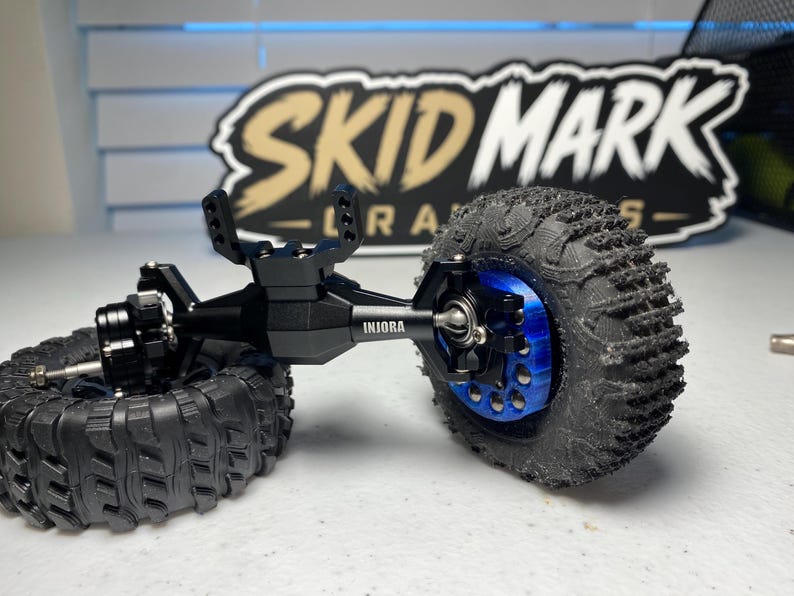 Op de afbeelding: Een zwarte metalen RC crawler as-assemblage met de merknaam INJORA in het wit gedrukt. De as is verbonden met twee zwarte rubberen banden met diep profiel, waarbij de rechterband op een felblauwe metalen wielnaaf is gemonteerd.