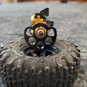 Op de afbeelding: Een zwarte wielnaaf met vijf spaken en een centrale metalen moer, direct boven een zwarte rubberen band met profiel geplaatst. Het geheel is voorzien van een goudkleurig mechanisch onderdeel dat aan de achterzijde uitsteekt en technische details toont.