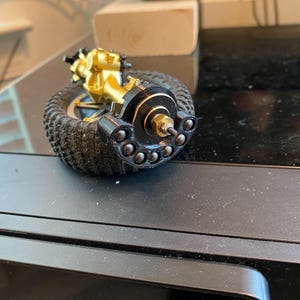 Op de afbeelding: Een goudkleurige metalen asconstructie voor een op afstand bestuurbare auto, voorzien van een zwarte ronde wielnaaf met een reeks kleine zilveren kogellagers in een halve cirkel, bevestigd aan een zwarte rubberen band met profiel.