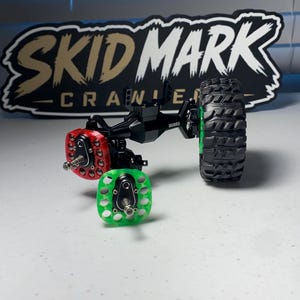 Op de afbeelding: Een zwarte metalen RC-crawler-asconstructie met een rode portaaltandwielafdekking, een groene portaaltandwielafdekking en &eacute;&eacute;n zwarte rubberen band met een diep profiel. De asbehuizing is voorzien van de merknaam INJORA in witte letters.
