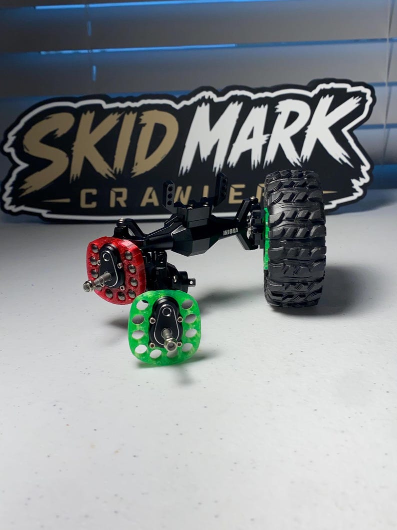 Op de afbeelding: Een zwarte metalen RC-crawler-asconstructie met een rode portaaltandwielafdekking, een groene portaaltandwielafdekking en &eacute;&eacute;n zwarte rubberen band met een diep profiel. De asbehuizing is voorzien van de merknaam INJORA in witte letters.