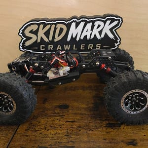 Puede incluir: Crawler a control remoto negro y plateado con neumáticos grandes y texturizados. El crawler tiene un chasis negro y componentes de suspensión rojos. El texto "SKID MARK CRAWLERS" se muestra encima del crawler.
