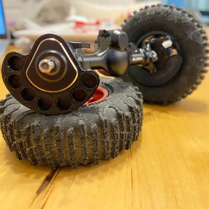 Op de afbeelding: Een zwarte metalen RC-crawler-asconstructie met een uniek cirkelvormig naafontwerp met meerdere gaten en een gouden rand, verbonden met een zwarte asbehuizing. Twee grote, getextureerde zwarte rubberen off-road banden met profiel zijn bevestigd.