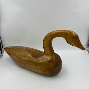 May include: A hand-carved wooden goose decoy featuring a smooth, polished light brown finish with visible natural wood grain patterns. The sculpture depicts a goose with an elongated neck, a curved head, and a pointed beak, showcasing craftsmanship.