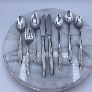 May include: A collection of vintage silverware, including spoons, forks, and knives, arranged on a round, marble-patterned surface. The silverware has a floral design on the handles and a shiny, reflective surface. The set includes six spoons, two knives, and one fork.