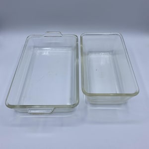 Peut inclure: Deux plats à four rectangulaires en verre transparent. L'un est plus grand avec des poignées, l'autre est plus petit. Les deux sont vides et semblent être en verre résistant à la chaleur, adapté à une utilisation au four. Les plats sont sur une surface blanche.
