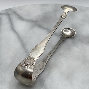 Pode incluir: Uma pinça de açúcar em metal prateado com um motivo decorativo de concha na dobradiça e ao longo dos cabos. A pinça possui duas extremidades arredondadas em forma de colher e um detalhe de borda frisada ao longo dos braços de metal.