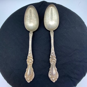 May include: Two silver-toned metal spoons featuring an ornate, embossed floral and scrollwork pattern along the handles. The spoons have a polished, reflective finish on the bowls and a detailed, textured design on the stems and base of the handles.