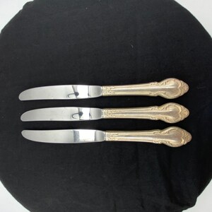 May include: Three silver-toned dinner knives with polished stainless steel blades and ornate, textured metal handles featuring a decorative scrollwork pattern at the base. The knives are arranged horizontally, showcasing a uniform design.