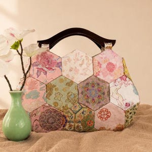 Puede incluir: Un bolso de mano construido con múltiples paneles de tela hexagonales que presentan varios patrones florales, geométricos y de animales en tonos rosa, verde y dorado. El bolso incluye un asa de madera curva de color marrón oscuro.