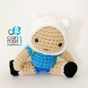 May include: A crocheted plush toy of Finn the Human from the animated series Adventure Time. Finn is wearing his signature white hat with pointed ears and a blue shirt. He is sitting with his legs crossed and has a friendly expression on his face.