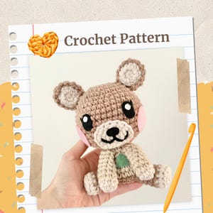 Puede incluir: Un oso de ganchillo con cuerpo marrón, orejas color crema y un triángulo verde en el pecho. El oso tiene ojos negros, mejillas rosas y nariz y boca negras. Las palabras "Crochet Pattern" están encima del oso.
